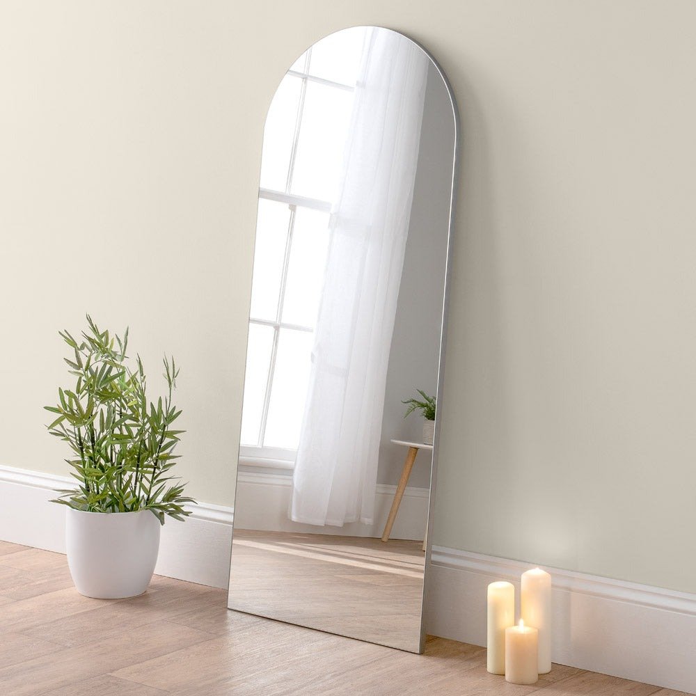 Full-length mirror leaning against a wall with a plant and candles on a wooden floor.