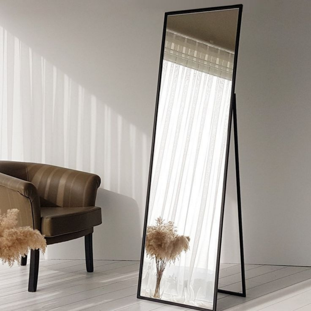 Full-length mirror with black frame leaning against a wall in a room with a brown chair and decorative plant.